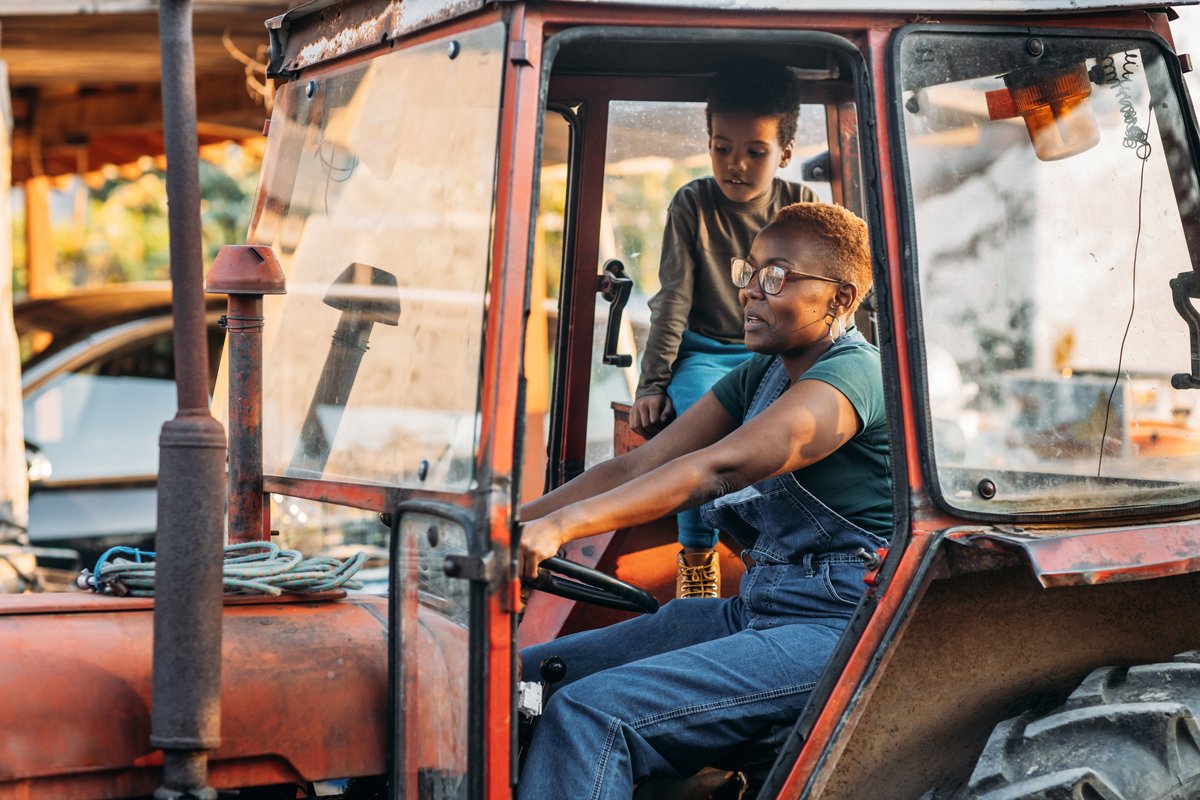 A Black farmer and a child drive a tractor on the farm.