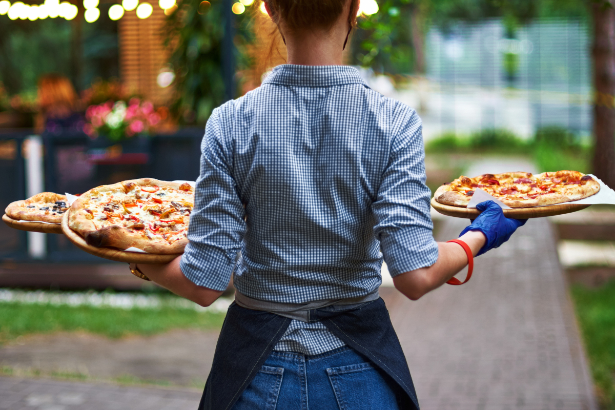 Female restaurant worker with her back to us with three pizzas in her hands on the restaurant terrace