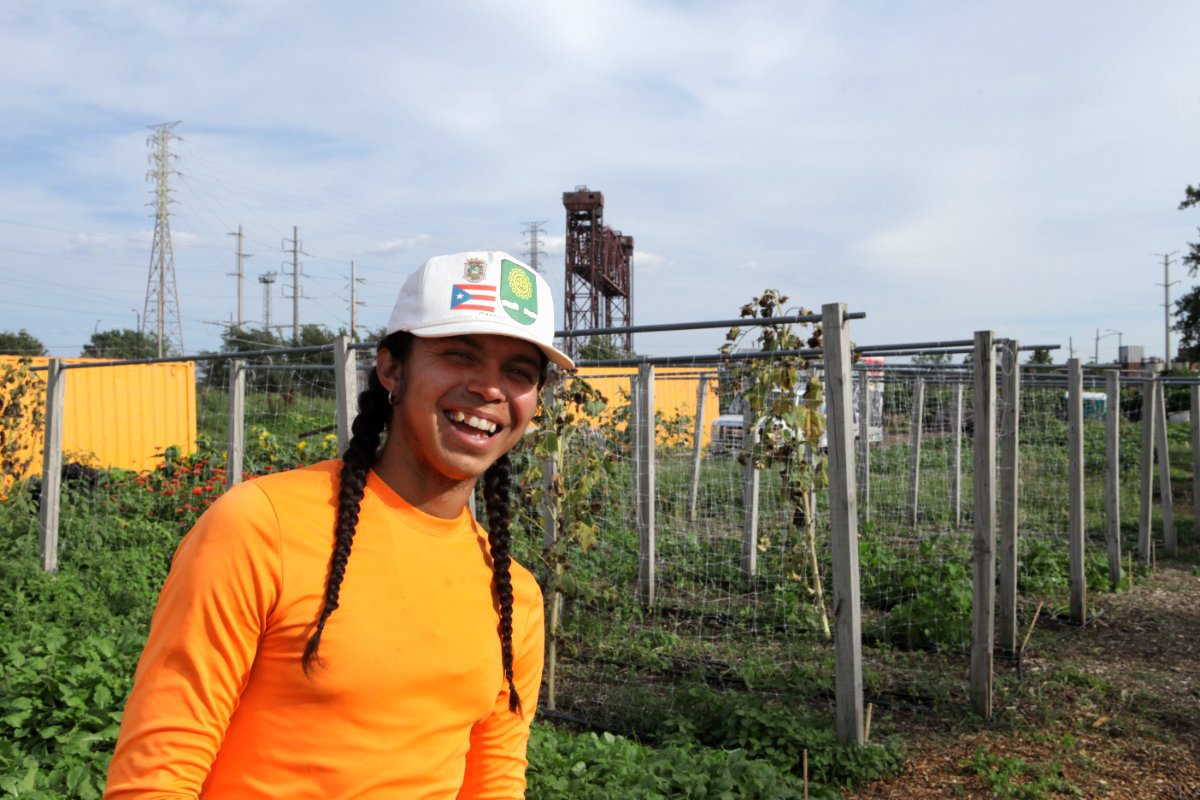 Xavier “X” Colon, a young person with long braided black hair, a baseball cap, and a bright orange hi-visibility shirt, stands in the fields at South Chicago Farm. (Photo by Paul Gordon)