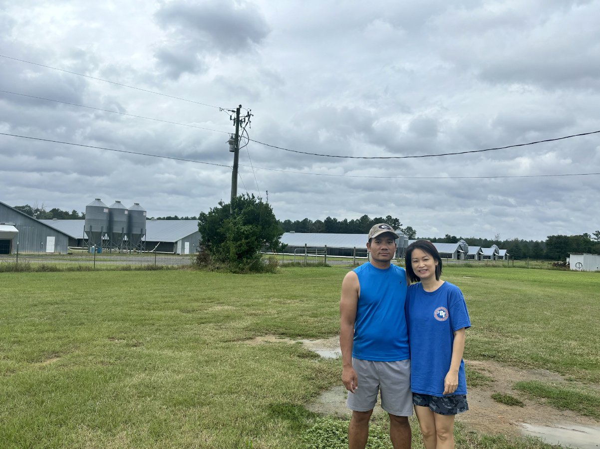 A Vietnamese American couple wearing blue t-shirts stands in a large green field on a chicken farm